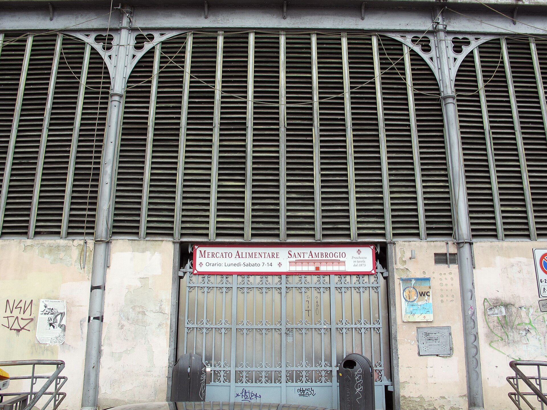 A Sant'Ambrogio market banner hangs above a closed iron gate beneath green louvres and decorated iron arches in Florence.