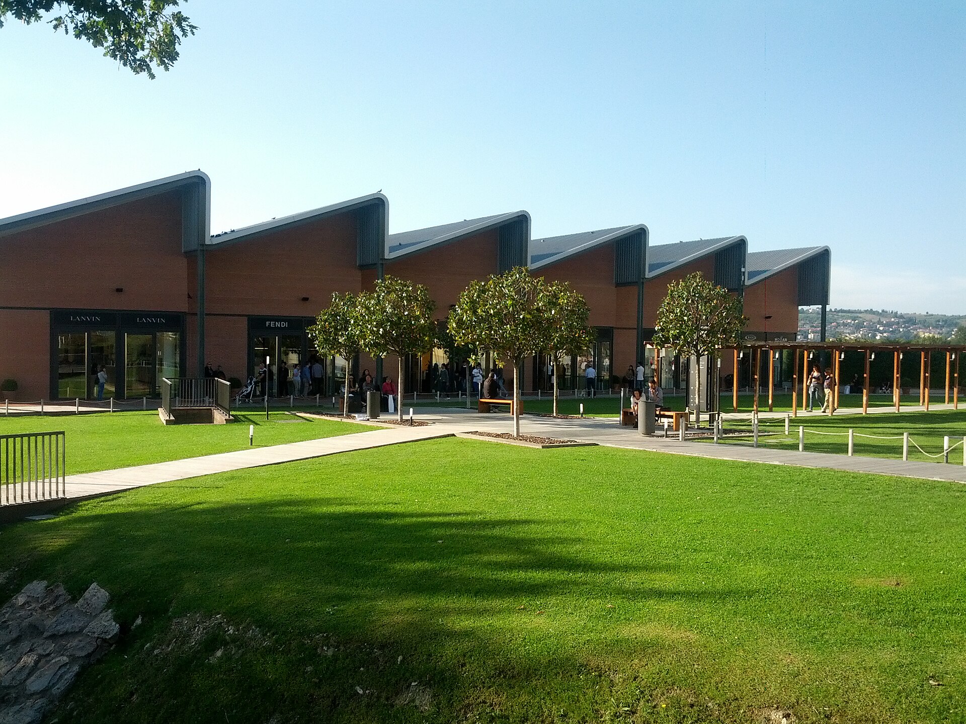 Sawtooth-roofed brick storefronts at The Mall outlet near Florence, with Lanvin and Fendi shops, a lawn in front and a few shoppers walking the paths