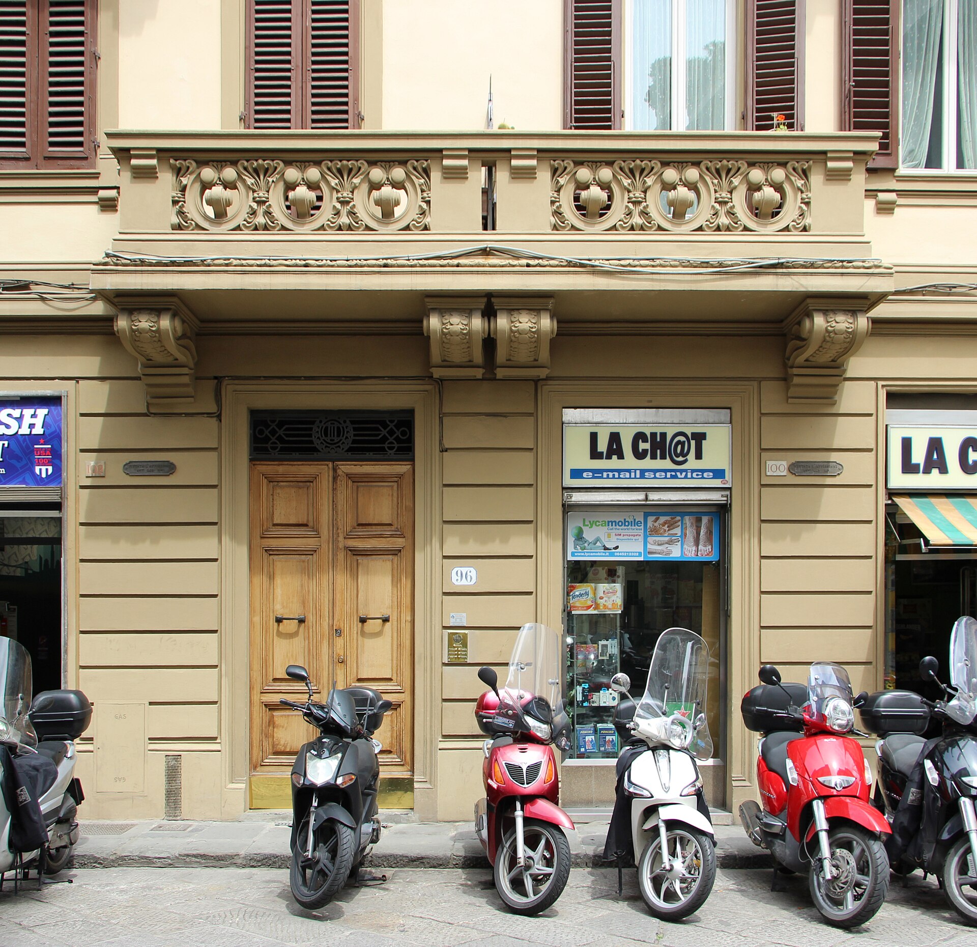 Scooters stand outside a wooden doorway beneath an ornate balcony on Via Ghibellina in Florence, beside a wall-mounted entry panel.