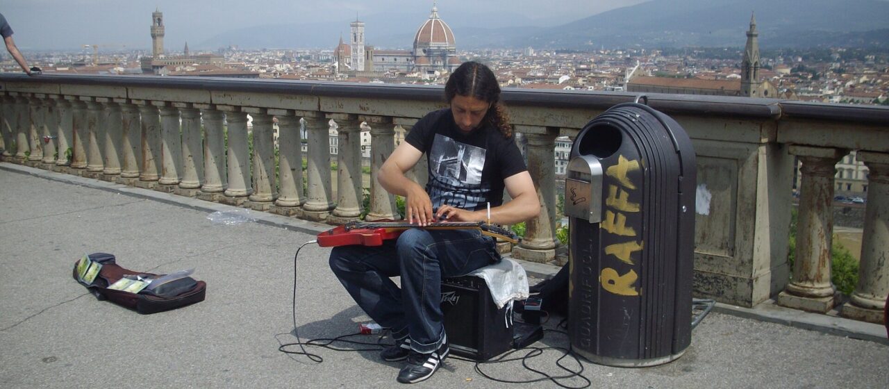 A seated street musician plays guitar at Piazzale Michelangelo above Florence, with the cathedral dome behind him.