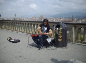 A seated street musician plays guitar at Piazzale Michelangelo above Florence, with the cathedral dome behind him.