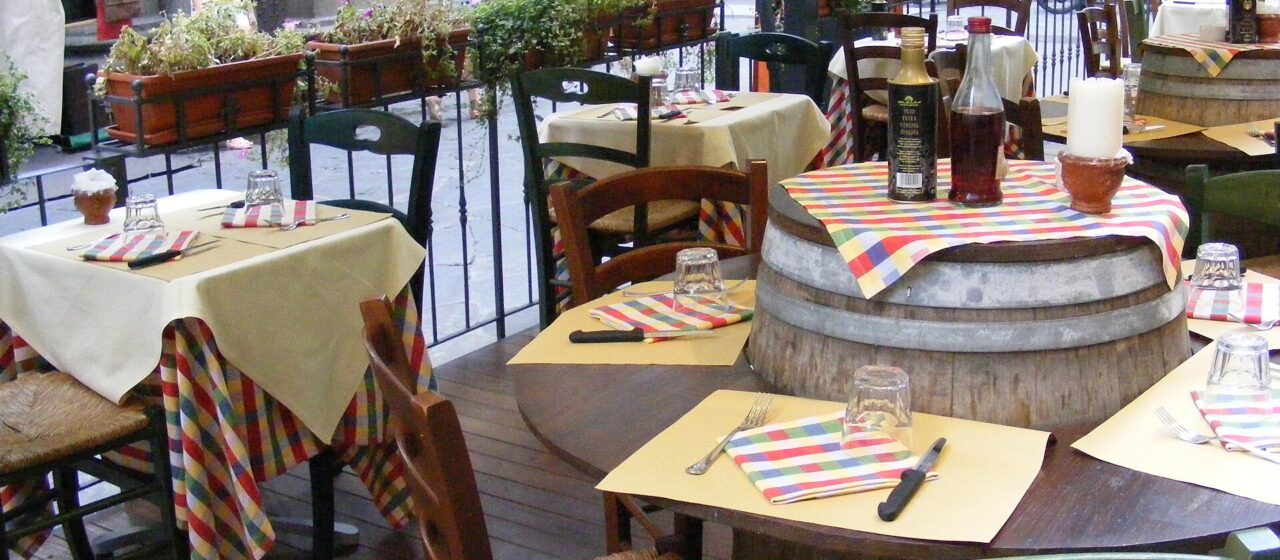 Set wooden tables, checked napkins and a barrel table line an outdoor Florence restaurant terrace beside street-side planters.