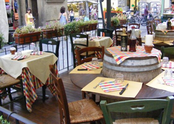 Set wooden tables, checked napkins and a barrel table line an outdoor Florence restaurant terrace beside street-side planters.