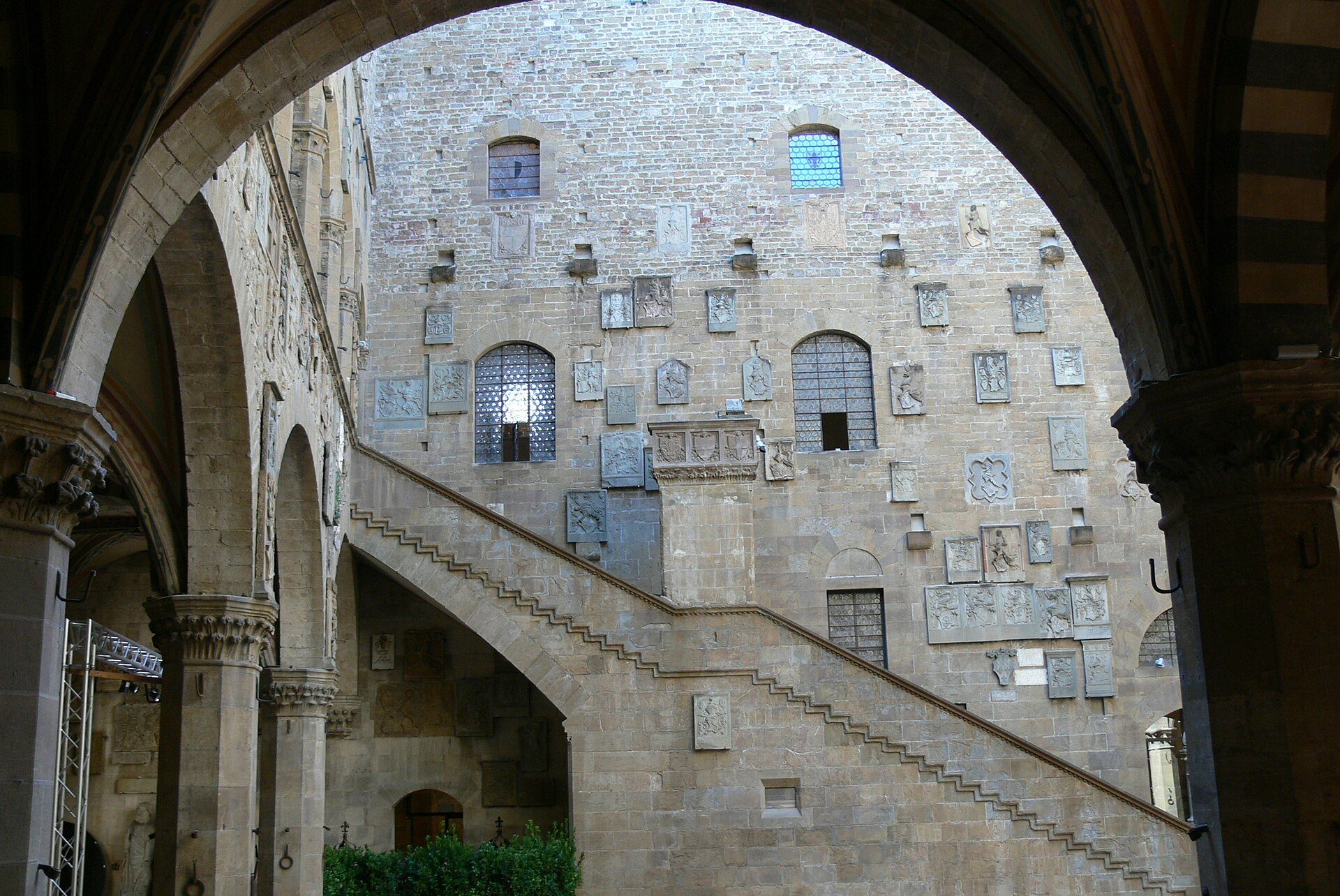 A shaded arch frames the outdoor staircase and walls covered with coats of arms in Florence's Bargello courtyard.