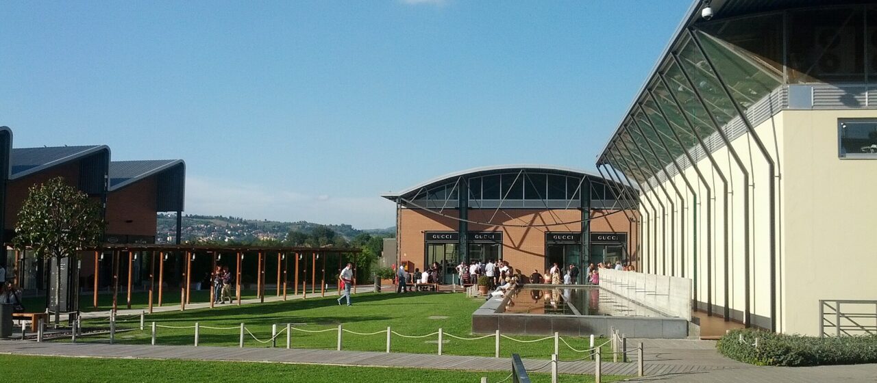 Shoppers walk between modern storefronts and green lawns at The Mall outlet in Reggello, Tuscany.