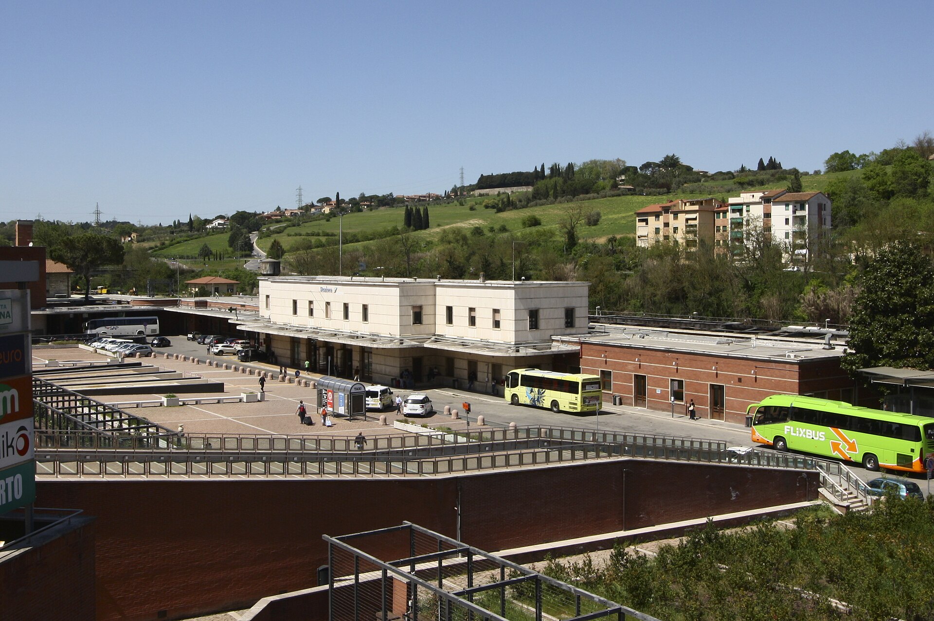 Siena railway station seen from above, a long low white building with a bus and coaches in the forecourt and green hills rising behind