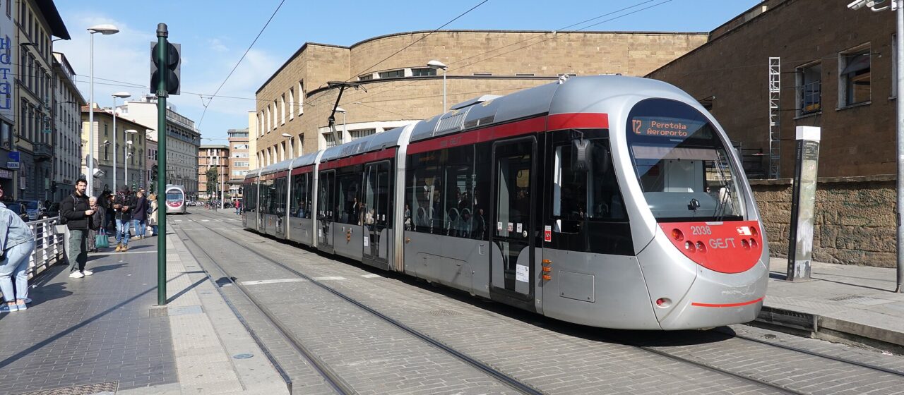A silver and red T2 tram passes Santa Maria Novella station in Florence, with pedestrians alongside the street tracks.