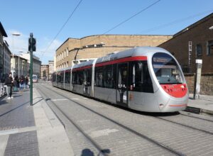 A silver and red T2 tram passes Santa Maria Novella station in Florence, with pedestrians alongside the street tracks.