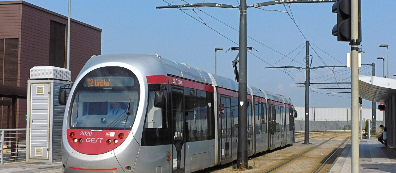 A silver and red T2 tram waits between the platforms at Florence Airport beneath overhead wires and blue sky.