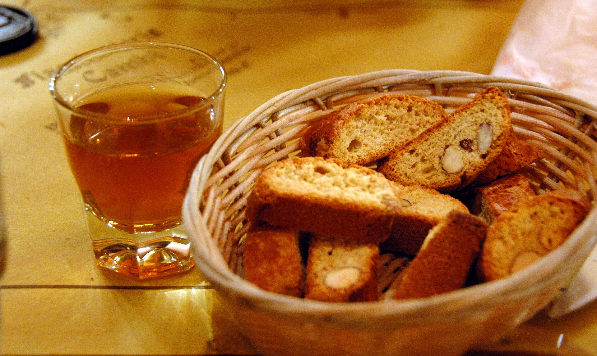 Sliced almond cantucci fill a wicker basket beside a small glass of amber vin santo on a wooden table.
