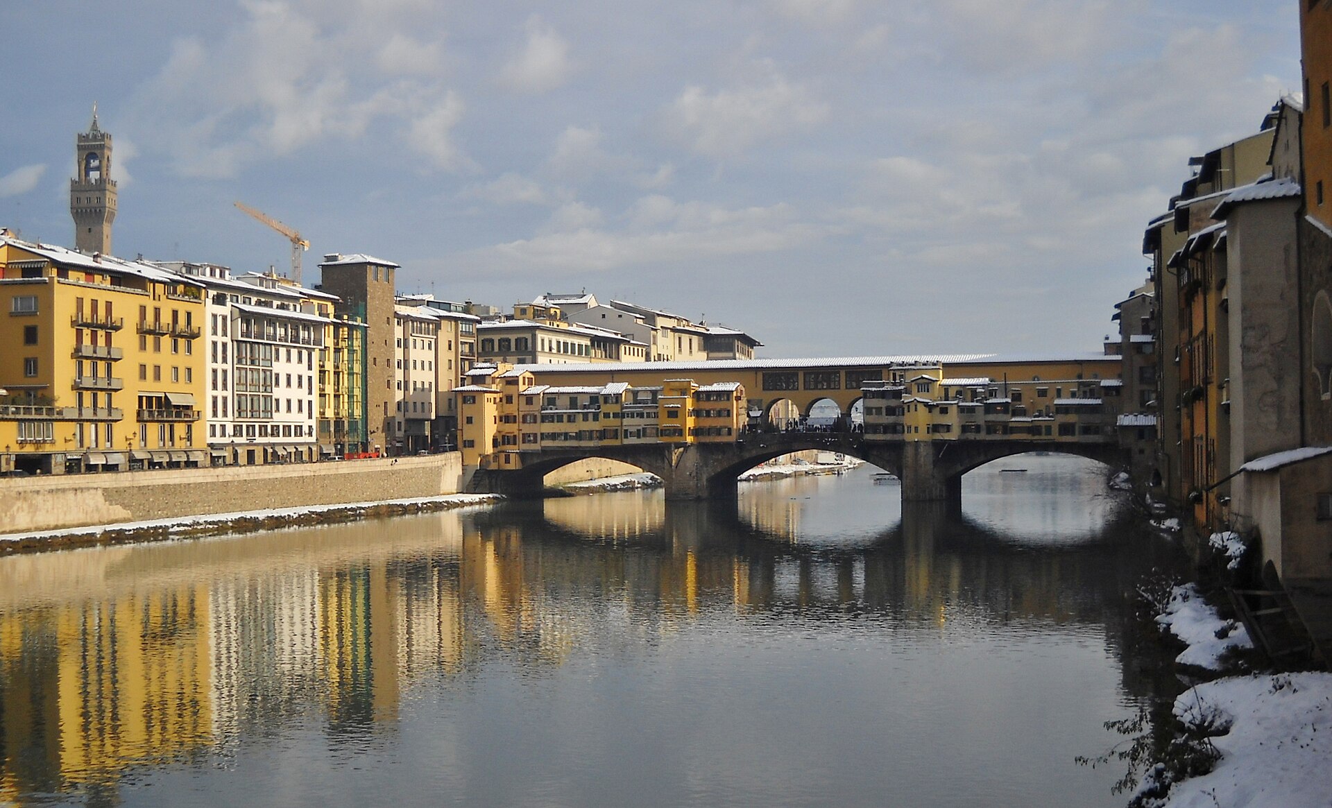 Snow rests on Florence’s riverside roofs and banks while sunlit Ponte Vecchio and yellow buildings reflect in the calm Arno.