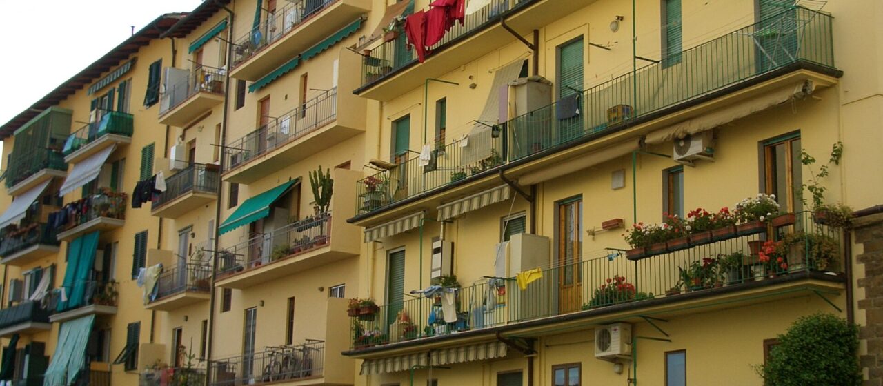 Stacked balconies, green shutters, flower boxes and drying laundry cover a yellow apartment building in Florence.