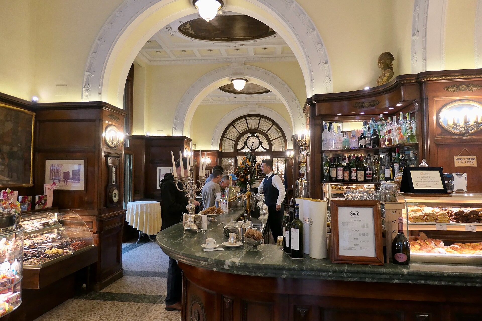 Staff and customers gather around Caffe Gilli's curved marble counter between pastry cases beneath cream arches in Florence.