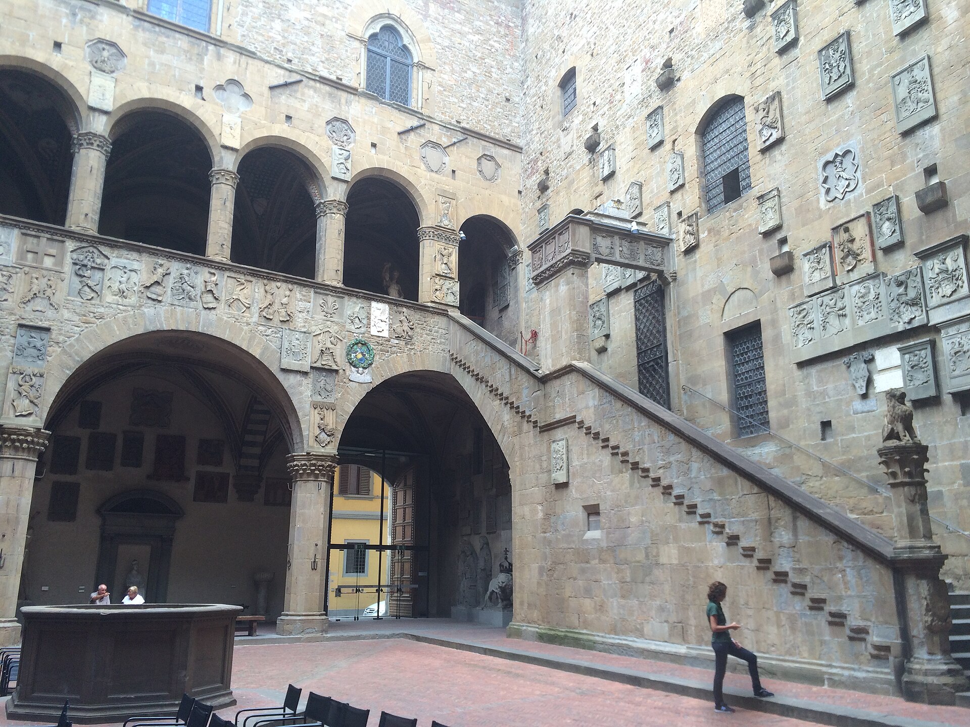 Stone stairs cross the heraldic courtyard of Florence's Bargello, beside an arcaded loggia and a well.
