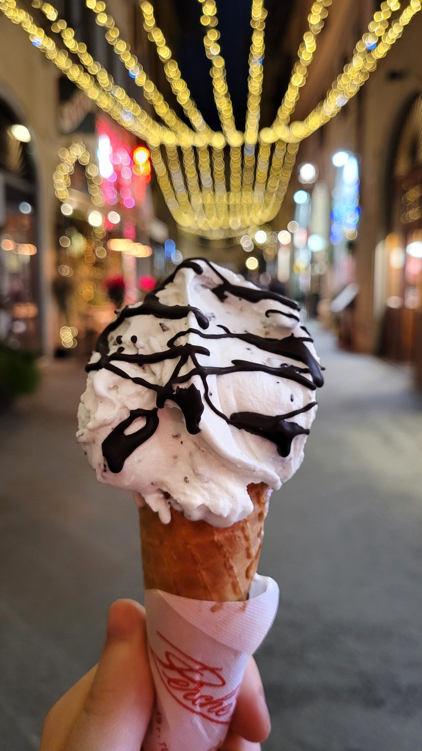 A stracciatella gelato cone topped with chocolate is held beneath strings of lights on Florence's Via dei Tavolini.