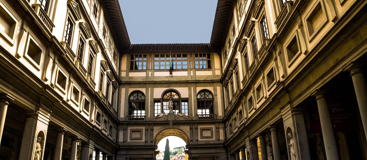 Sunlight catches the Uffizi's upper windows above visitors gathered between its two wings in the Piazzale degli Uffizi, Florence.
