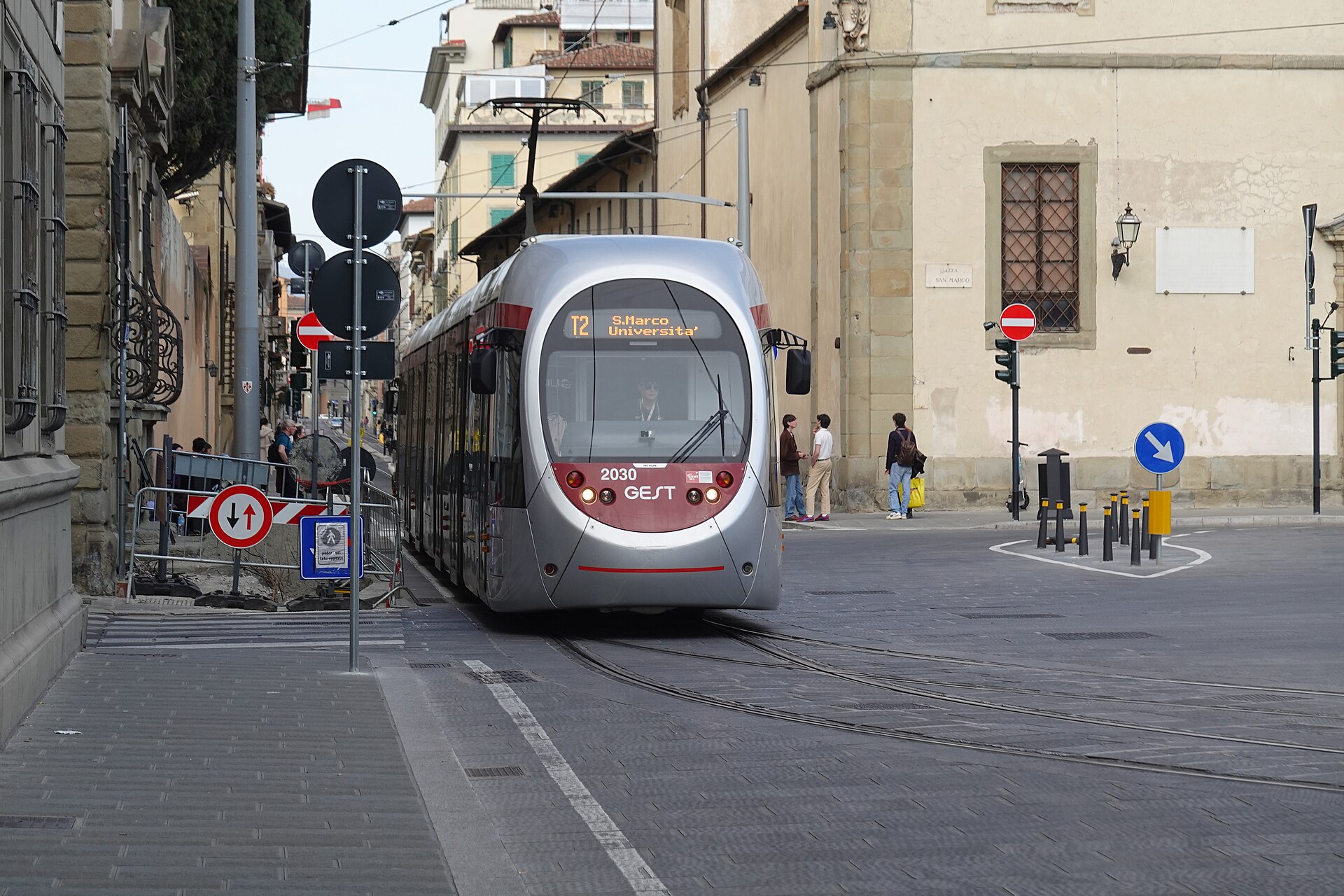 A T2 tram runs between pale stone buildings on a narrow Florence street, with pedestrians beside the tracks.