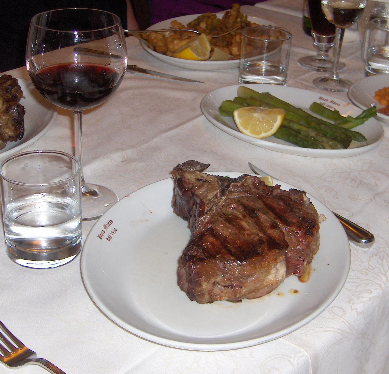 A thick grilled steak sits on a white plate beside red wine, asparagus and side dishes at a Florence dinner table.