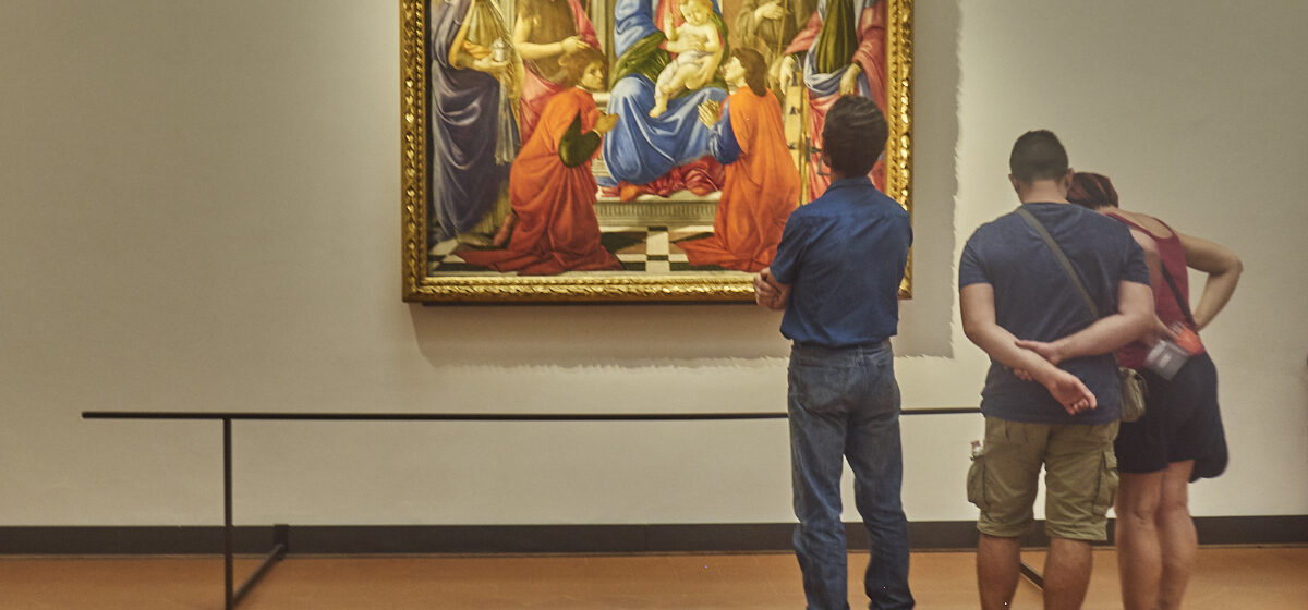 Three visitors study a gilded Madonna and Child painting with saints inside a gallery room at the Uffizi in Florence.