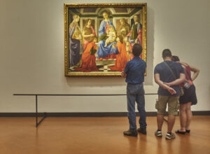 Three visitors study a gilded Madonna and Child painting with saints inside a gallery room at the Uffizi in Florence.
