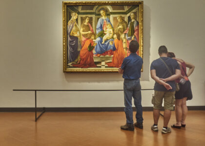 Three visitors study a gilded Madonna and Child painting with saints inside a gallery room at the Uffizi in Florence.