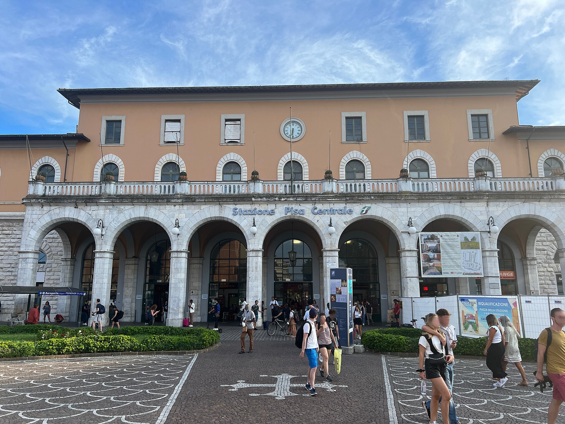 Travelers cross the paved forecourt of Pisa Centrale station beneath its clock and arched entrance arcade.