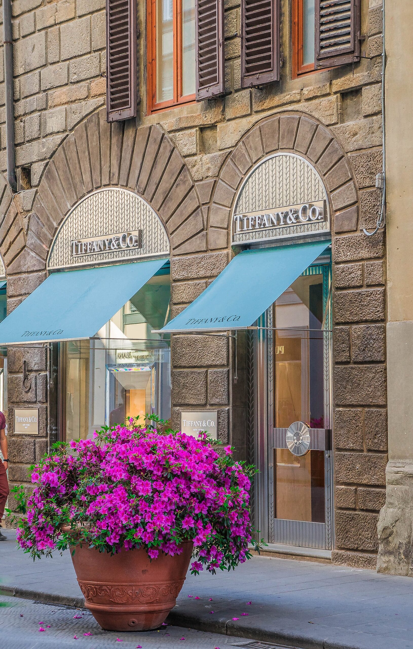 Turquoise Tiffany awnings shade arched shop windows beside pink flowers on Florence's Via de' Tornabuoni.