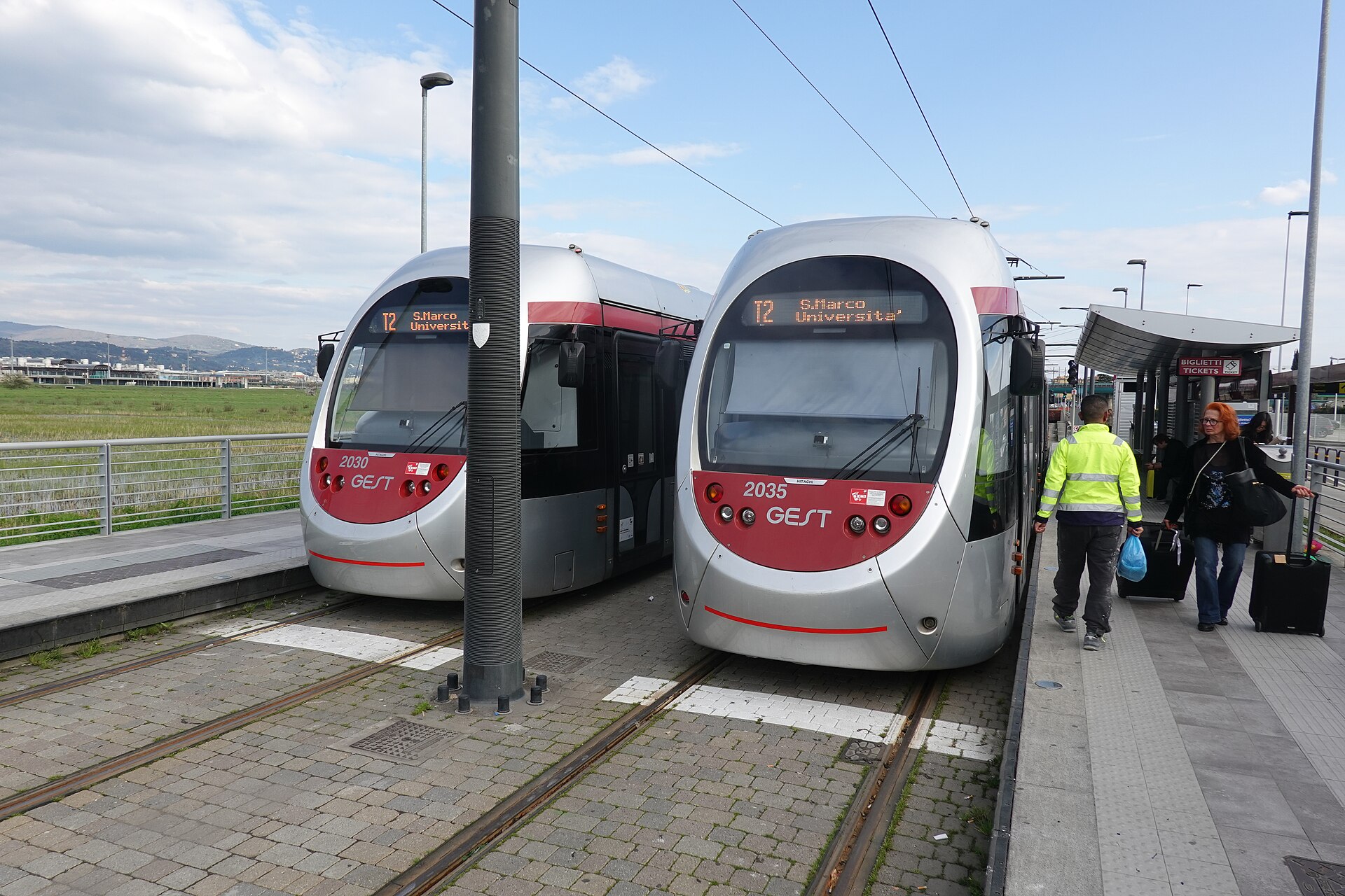 Two silver and red trams wait beside travelers with luggage at Florence Airport’s outdoor tram platforms.