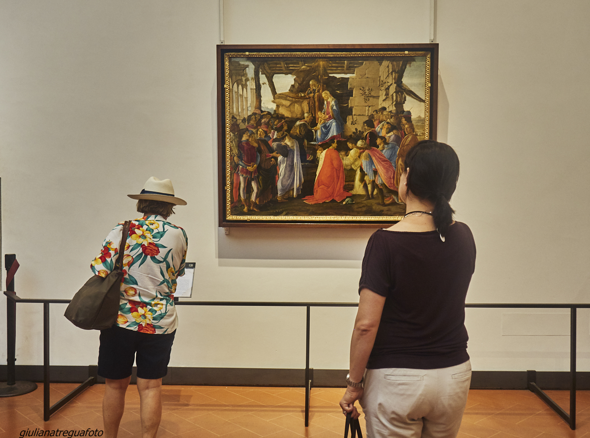 Two visitors look at a framed Renaissance religious painting with ruined architecture on a pale wall inside Florence's Uffizi Gallery.