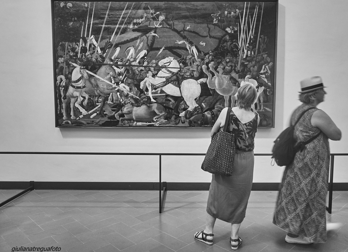 Two visitors stand before a large Renaissance battle painting with horses and lances in Florence's Uffizi Gallery.