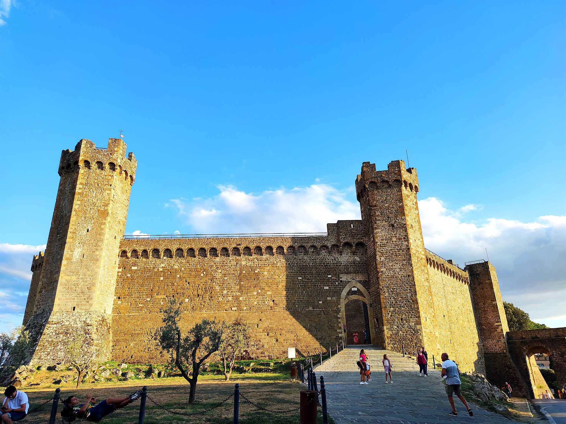 Visitors approach Montalcino’s sunlit stone fortress between crenellated towers, with lawns and a small tree beside the entrance.