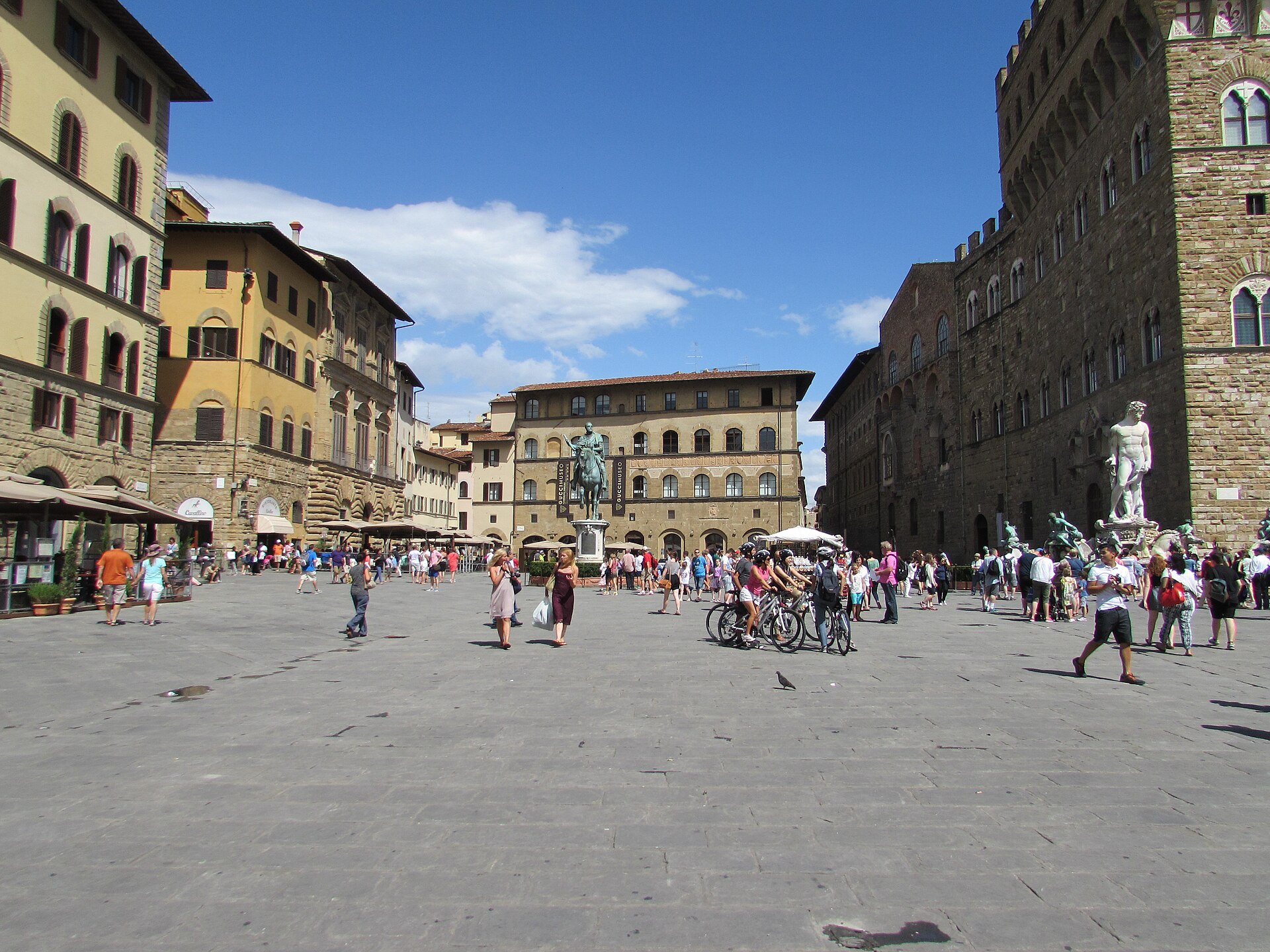 Visitors and cyclists cross Florence’s Piazza della Signoria beside Palazzo Vecchio and the equestrian statue of Cosimo I.