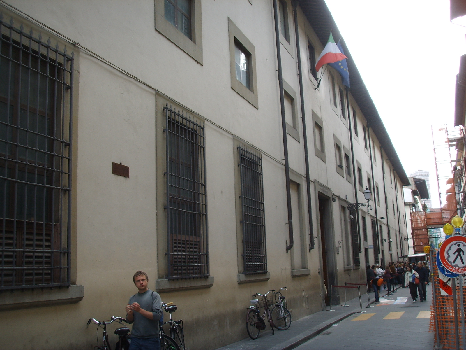 Visitors gather along the narrow street outside Florence's Accademia Gallery, beside its barred windows, entrance doorway and barrier posts.