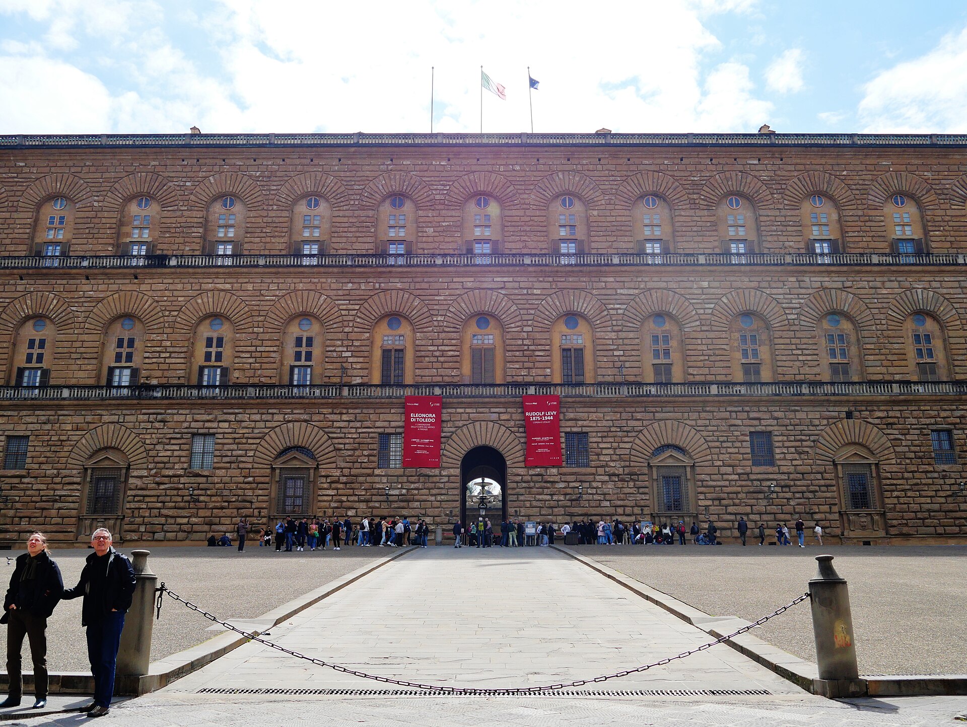 Visitors gather around the central arched entrance of Palazzo Pitti in Florence, beneath its broad rusticated stone facade.