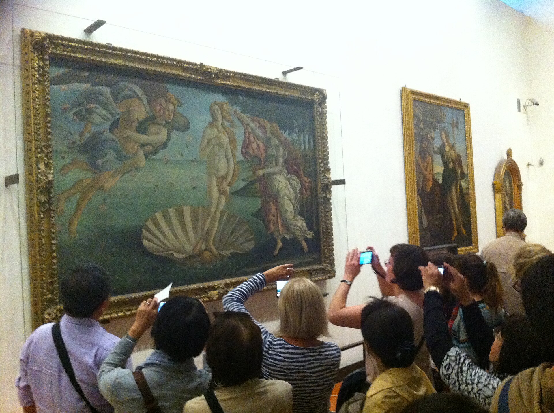 Visitors gather before Botticelli's framed Birth of Venus in Florence's Uffizi Gallery, with several holding cameras or phones.