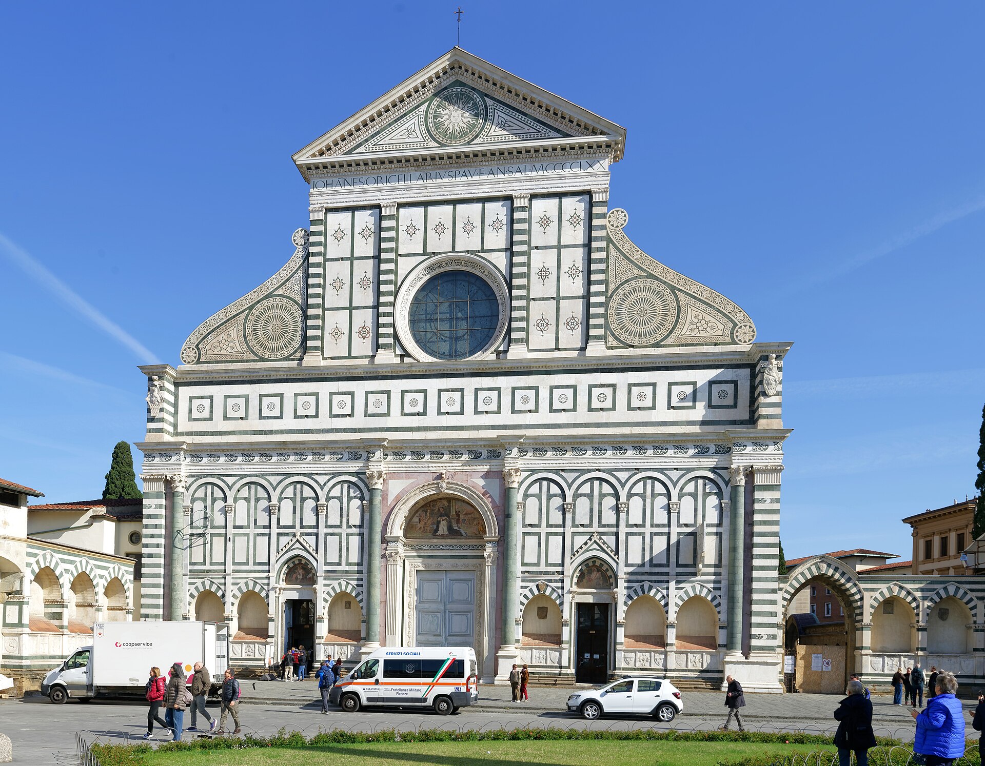 Visitors gather at Santa Maria Novella’s open side doorways in Florence while pedestrians pass parked vehicles in front of the church.