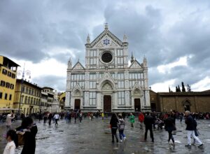 Visitors in jackets and long trousers gather by Santa Croce’s entrance steps and cross the wet piazza in Florence.
