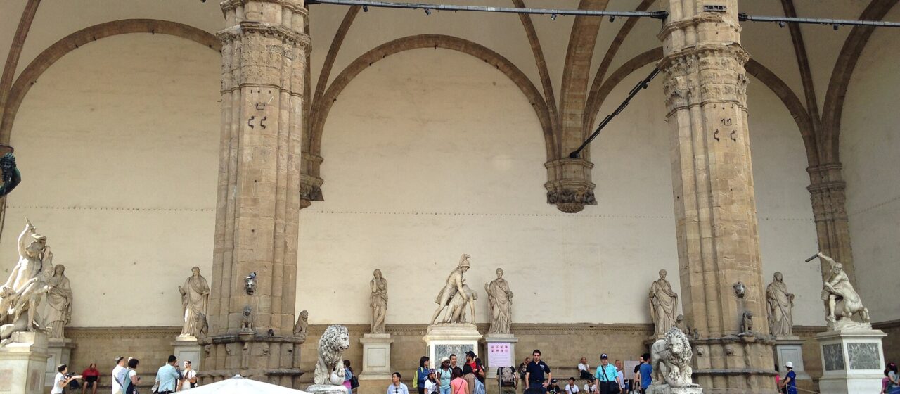 Visitors in light clothing gather and rest beside Florence’s Loggia dei Lanzi beneath its vaulted arches and marble sculptures.