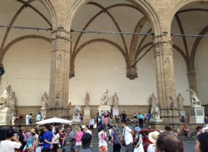 Visitors in light clothing gather and rest beside Florence’s Loggia dei Lanzi beneath its vaulted arches and marble sculptures.