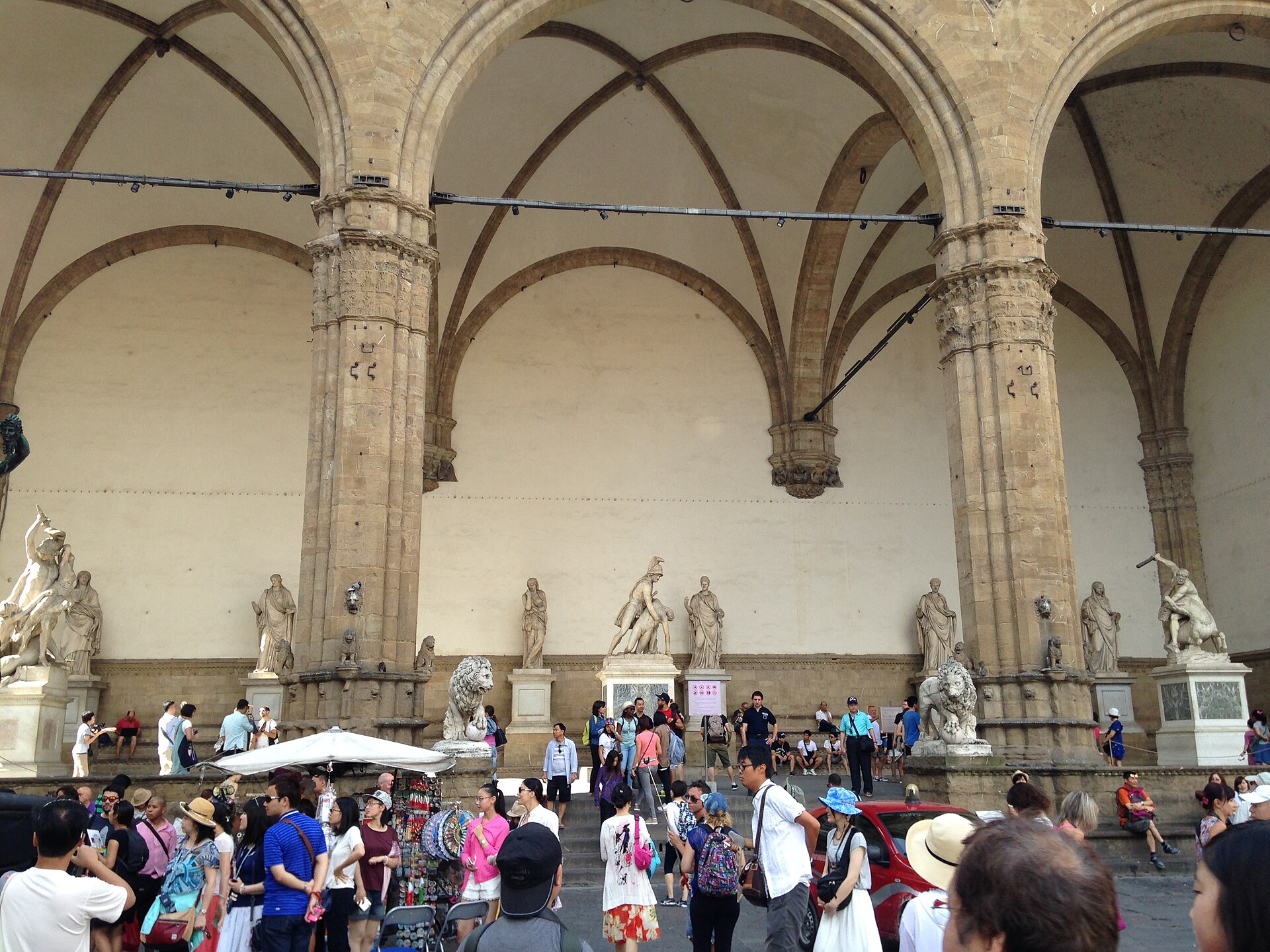 Visitors in light clothing gather and rest beside Florence’s Loggia dei Lanzi beneath its vaulted arches and marble sculptures.
