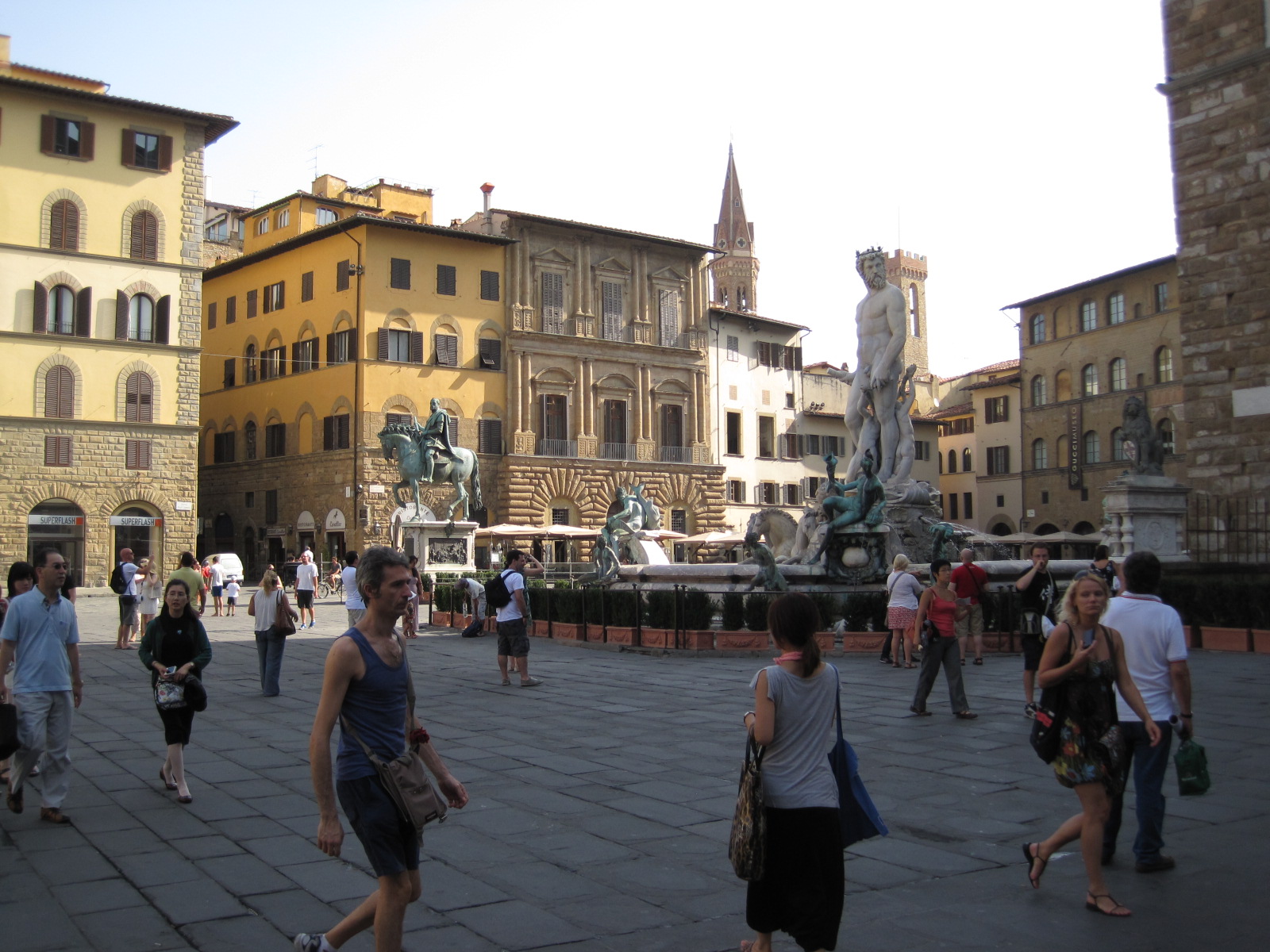 Visitors in light summer clothing walk beside the Neptune fountain in Florence’s Piazza della Signoria, with sunlit buildings behind.