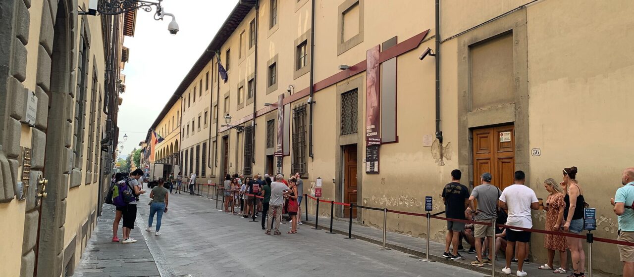 Visitors queue behind red rope barriers along the Accademia Gallery's facade on a narrow street in Florence.