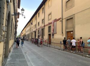 Visitors queue behind red rope barriers along the Accademia Gallery's facade on a narrow street in Florence.