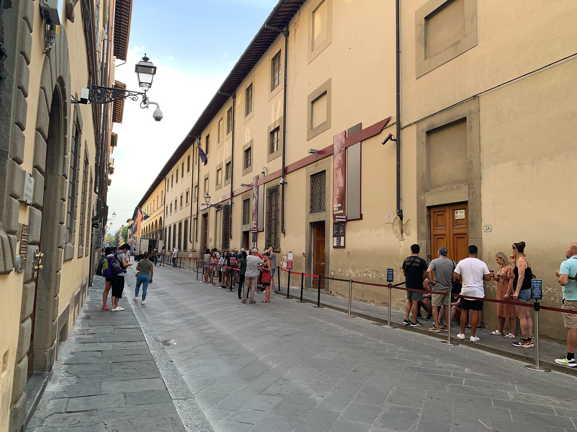 Visitors queue behind red rope barriers along the Accademia Gallery's facade on a narrow street in Florence.