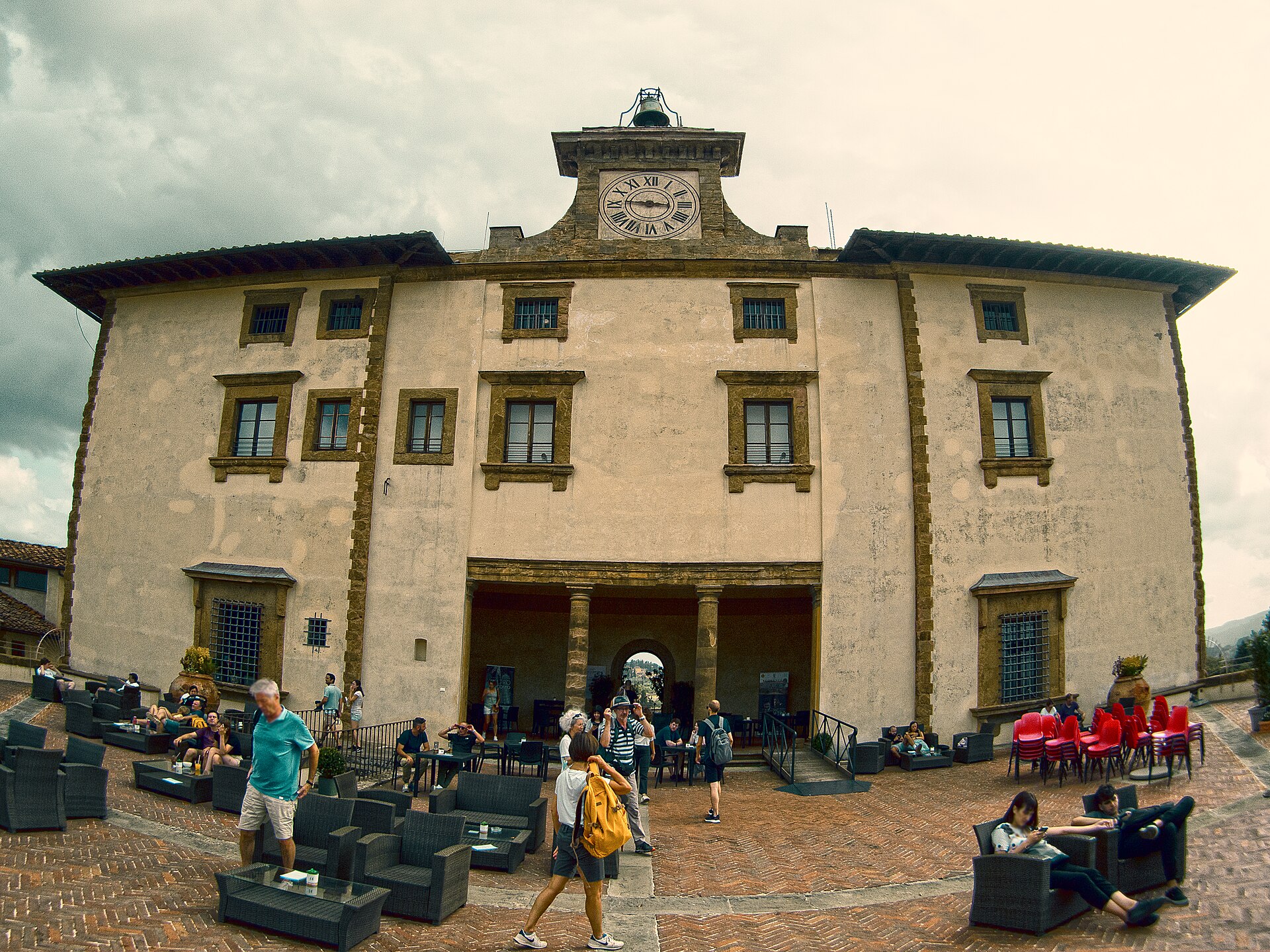 Visitors sit on lounge seating and walk across the brick terrace in front of the clock-towered Palazzina at Forte Belvedere in Florence, under a cloudy sky