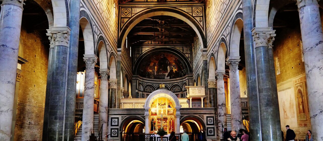 Visitors stand among stone columns beneath striped arches and a timber roof inside Florence's San Miniato al Monte.