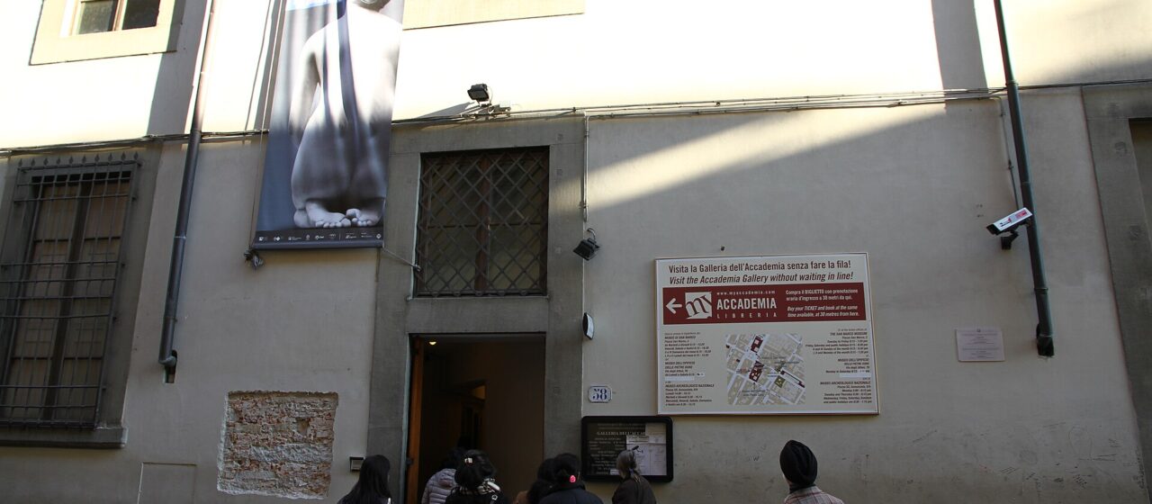 Visitors stand at the open entrance of Florence's Accademia Gallery, beneath an exhibition banner on its pale street facade.