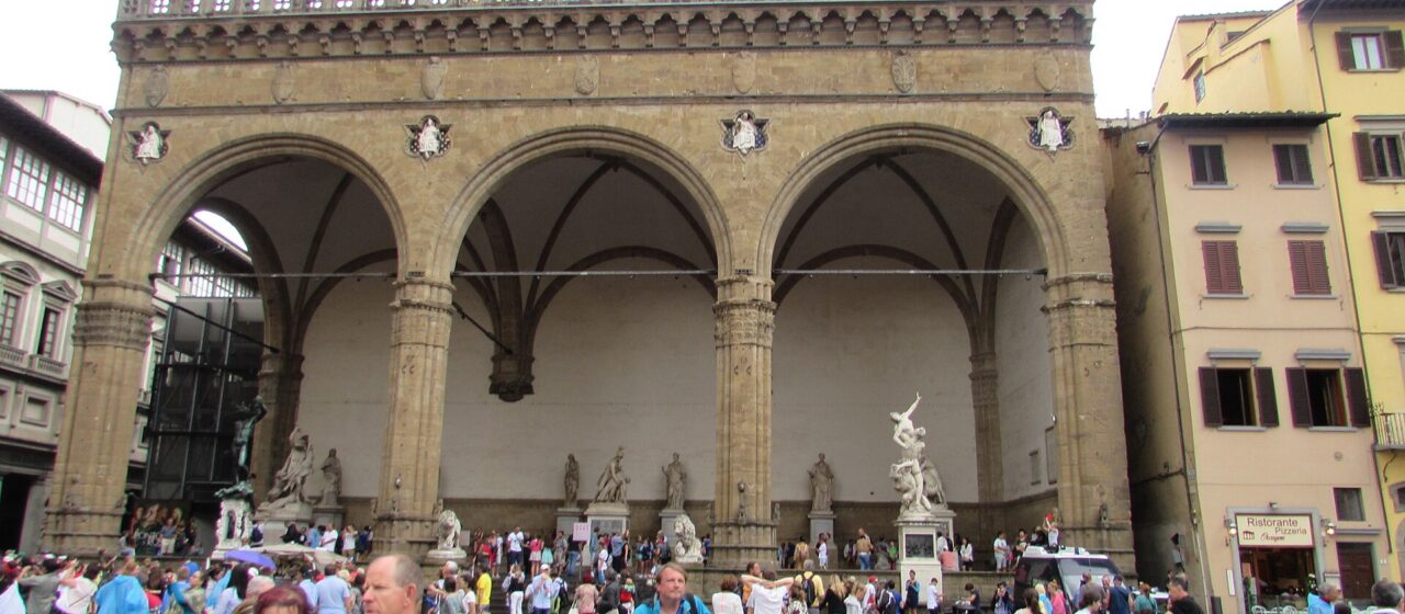 Visitors in summer clothes cross wet paving outside Florence’s Loggia dei Lanzi, with others resting beneath its three arches.