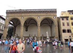 Visitors in summer clothes cross wet paving outside Florence’s Loggia dei Lanzi, with others resting beneath its three arches.