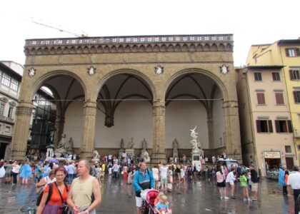 Visitors in summer clothes cross wet paving outside Florence’s Loggia dei Lanzi, with others resting beneath its three arches.